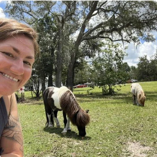 Owner smiling with two mini ponies grazing in a lush green field under shady oak trees.